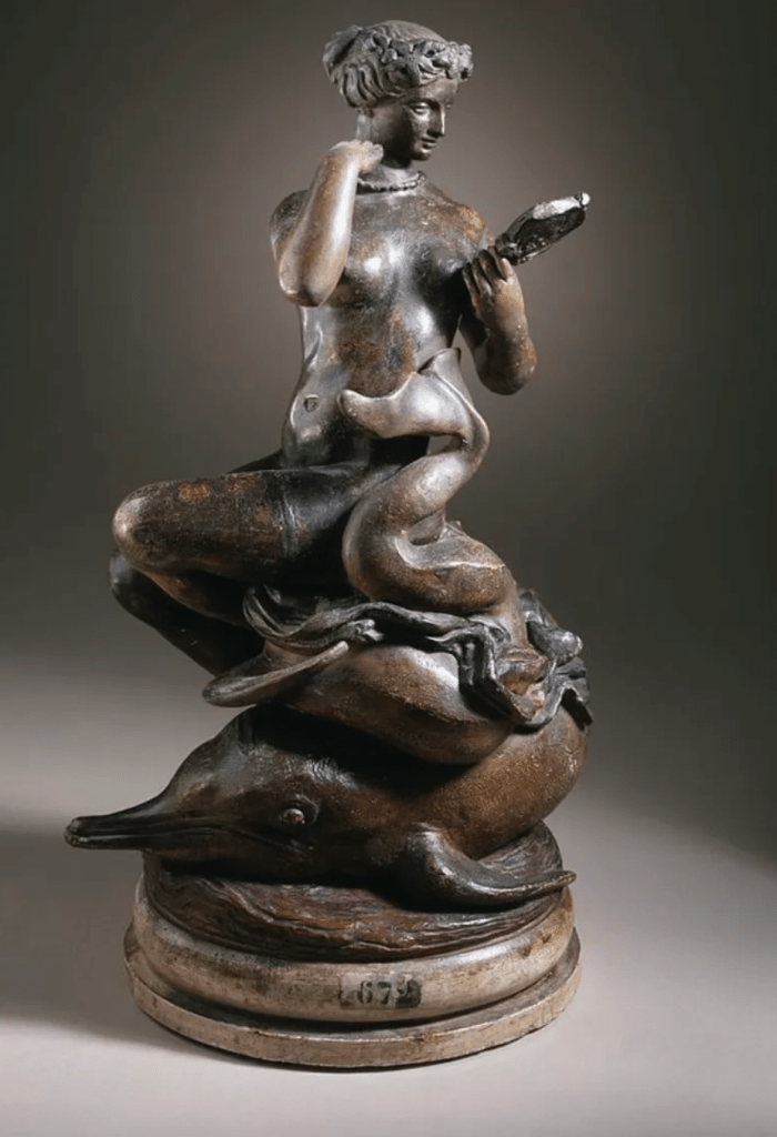 A small sculpted figure of a sea-nymph seated on a dolphin, arranging a necklace while rising from stylized waves.